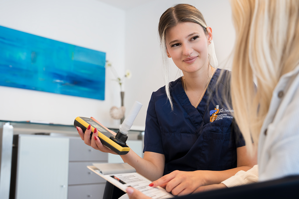 Eine medizinische Mitarbeiterin hält einer Patientin ein Atemtestgerät hin und zeigt auf ein Informationsblatt auf einem Klemmbrett