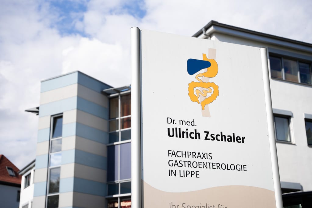 Blick von außen auf das Gebäude mit dem Praxisschild im Vordergrund.